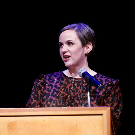 Photo of a woman with short hair wearing a leopard-print shirt speaking behind a podium with a microphone 
